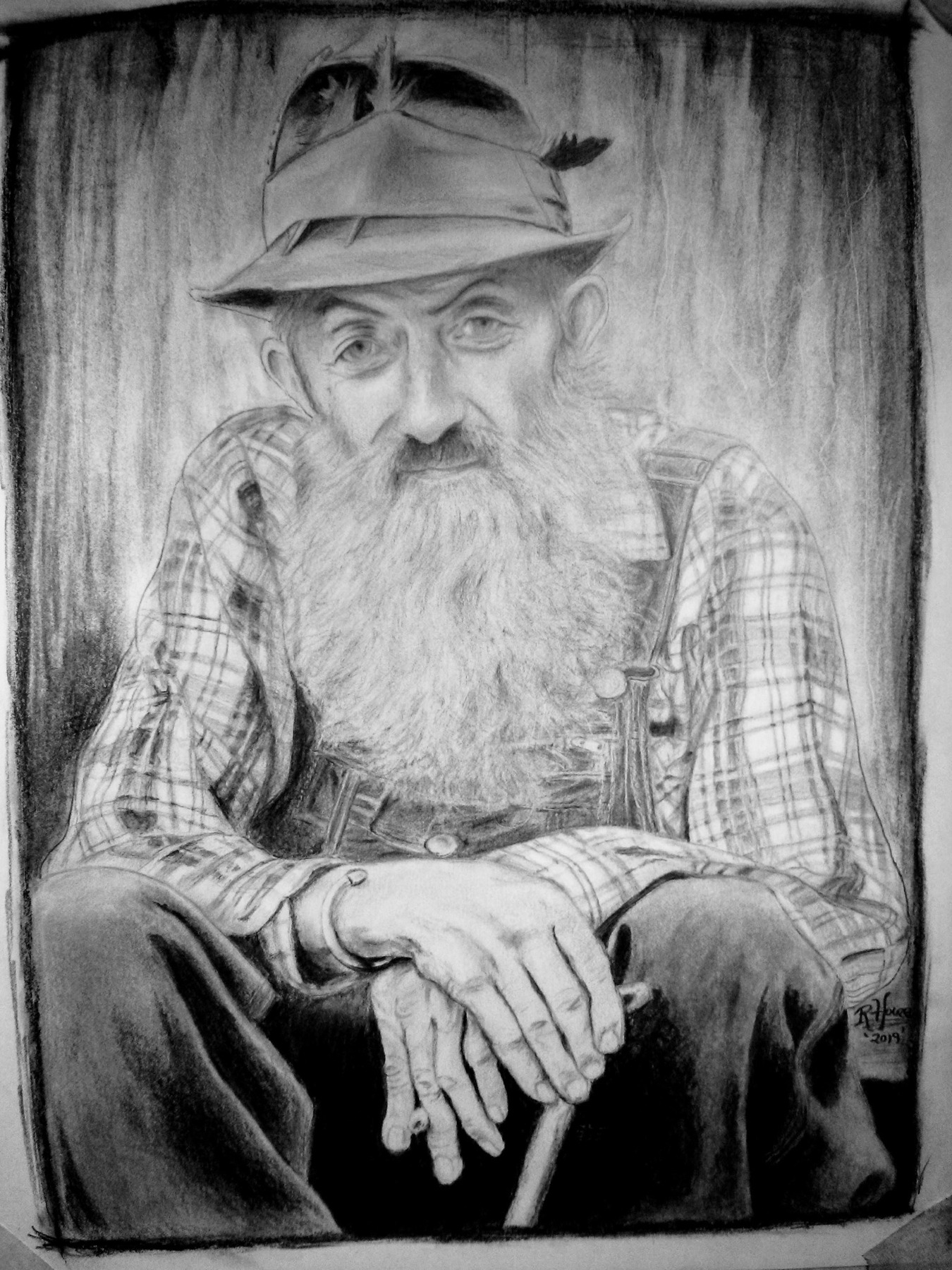 Seated bearded man in a hat, with overlapping hands and a checked shirt.